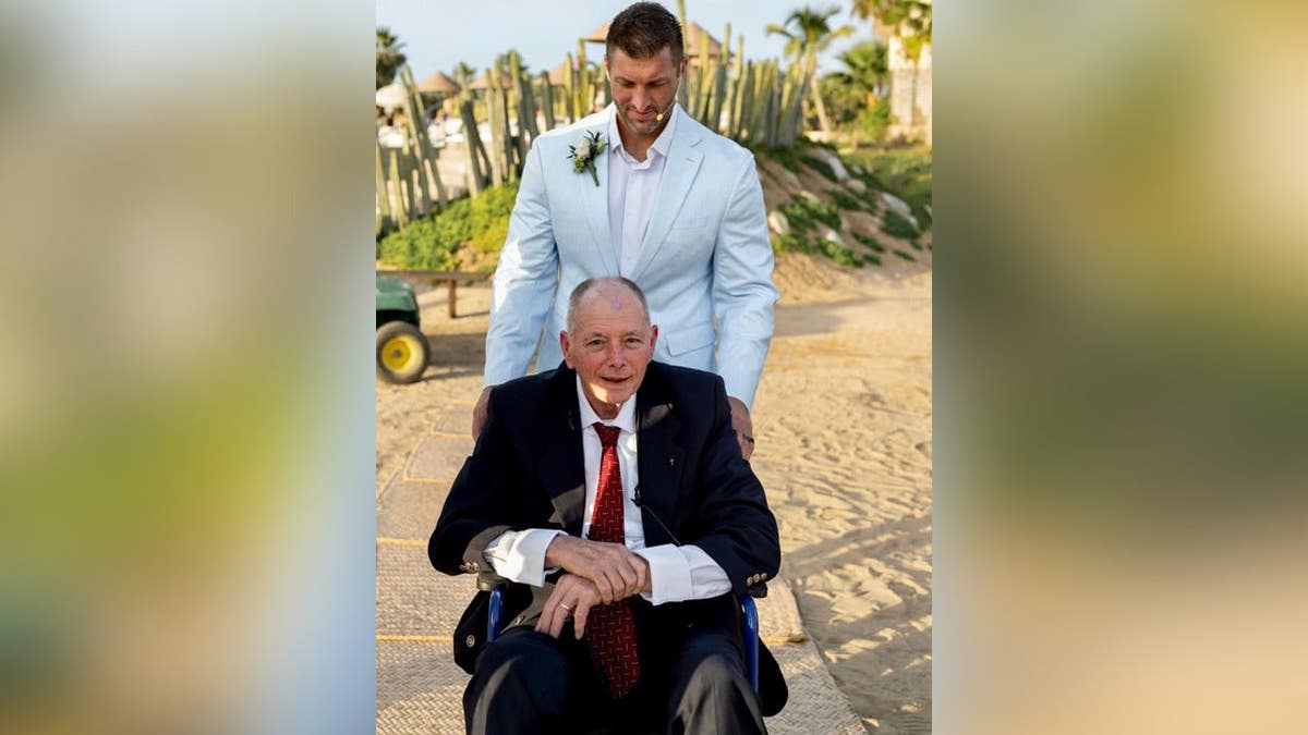 Tim Tebow seen pushing his father in wheelchair at wedding on beach.