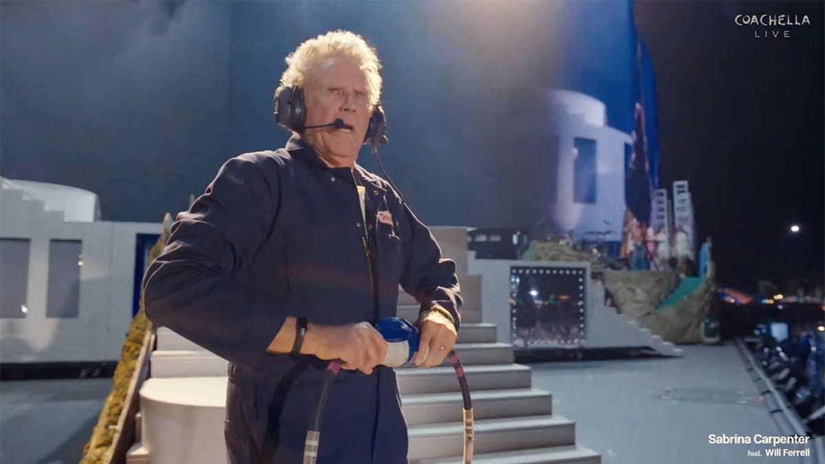 Will Ferrell in a jumpsuit holding a hose on a large set with steps and props, looking surprised.