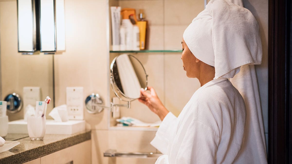 Young woman wearing a robe and towel on her head holding a mirror in a bathroom