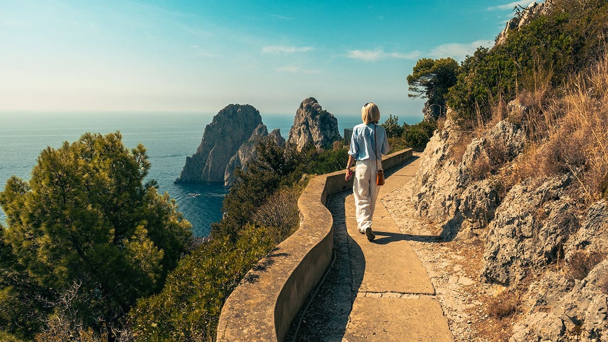 Woman walking along a coastal path in Capri with Faraglioni rock formations and sea view.