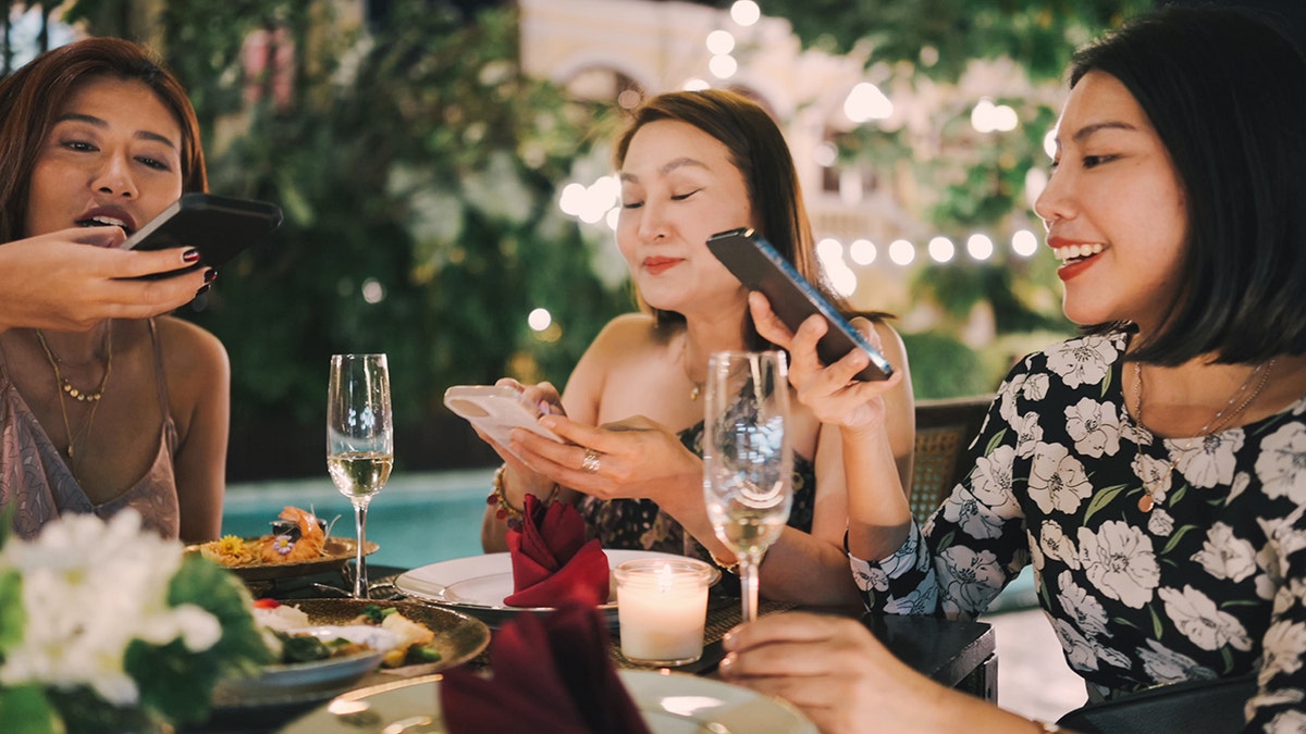 Women interact through mobile phones while dining, highlighting how social experiences merge with digital sharing modern social behavior.