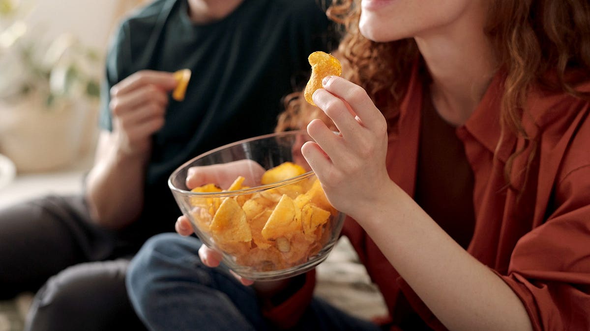 young woman holding a bowl of potato chips sitting in front of her boyfriend
