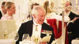 King Charles in a suit with medals smiling while sitting down at a state dinner.