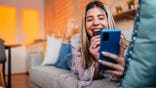 Young woman lying on sofa eating cookies and using smartphone