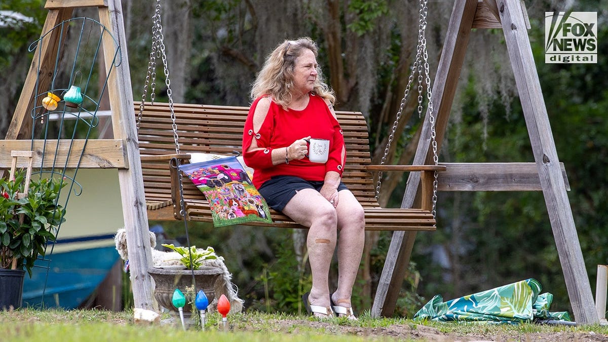 Becky Hill has a large coffee mug, a red shirt and denim jeans as she sits on a wooden bench swing outside her South Carolina home with a dog and boat in background