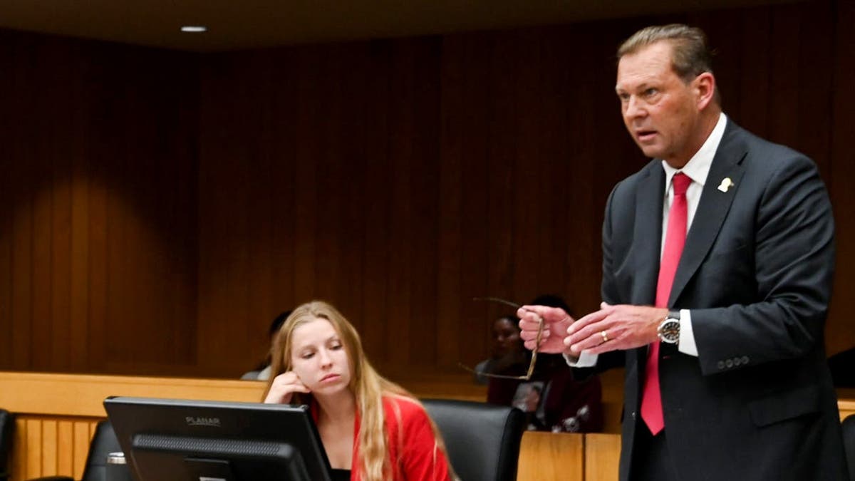 Doug Lloyd stands and speaks while Ken Fletcher and Andrea Marti sit at a table in a courtroom.