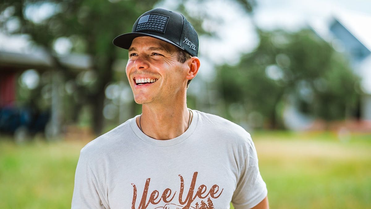 Granger Smith wearing a gray shirt and smiling as he sits in his truck and looks away from the camera.