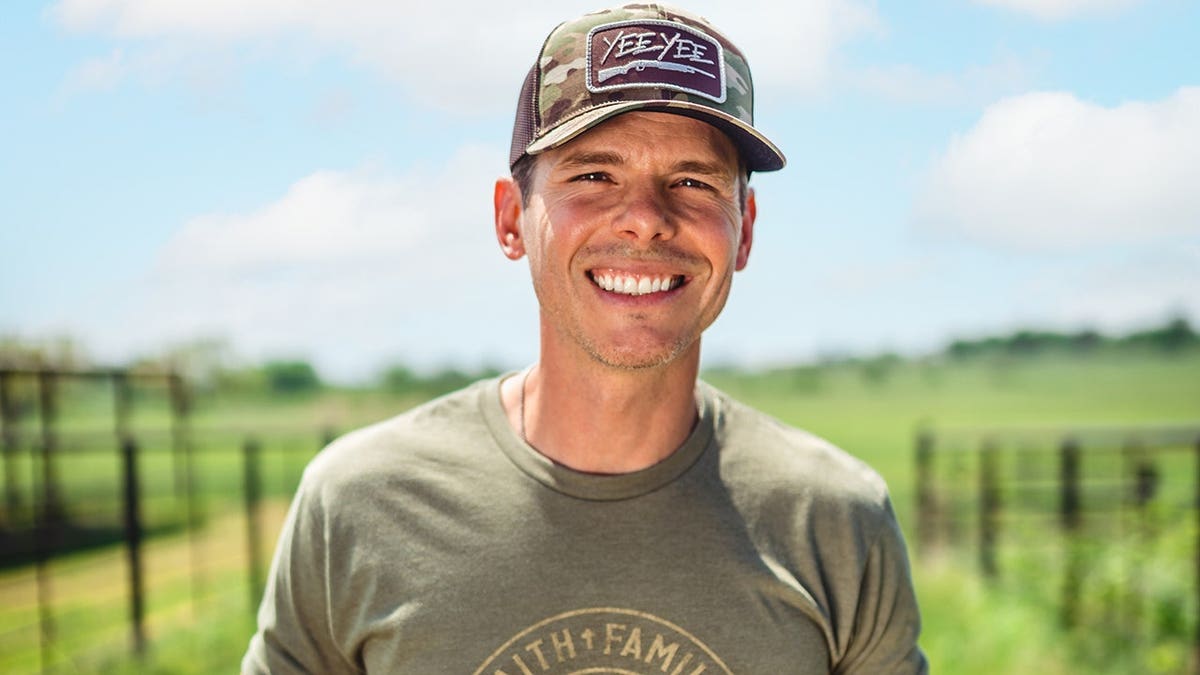 Granger Smith wearing a dark green shirt and smiling in front of the green field.