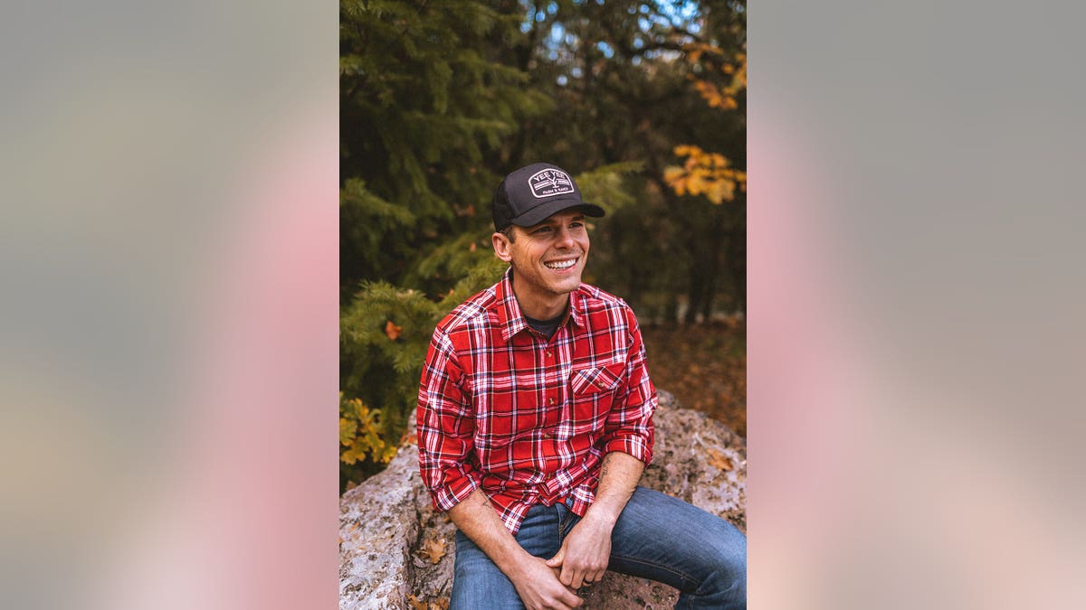 Granger Smith in a red plaid shirt and jeans sitting on a rock and smiling.