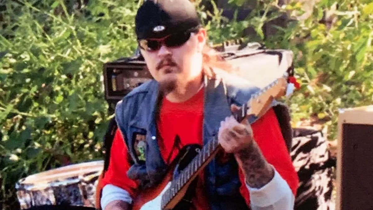 Keith Palumbo, con una camiseta roja y un chaleco vaquero, tocando la guitarra al aire libre.