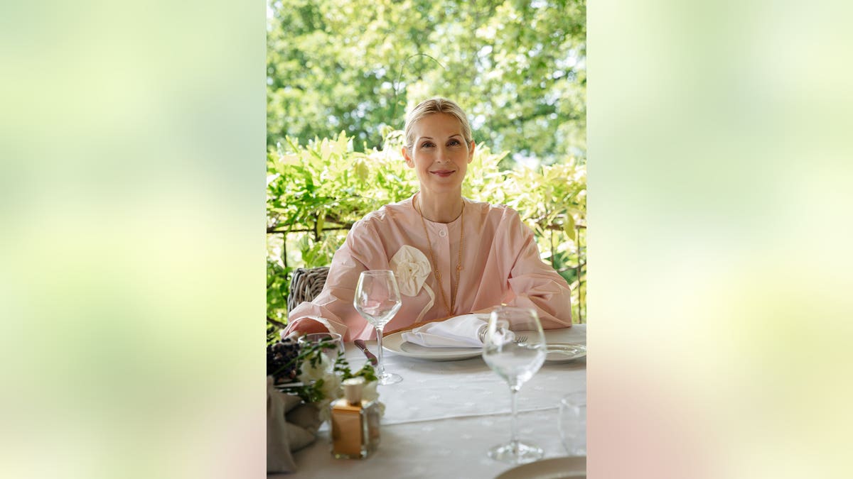 Kelly Rutherford sitting at a white table wearing a pink sweater next to her perfume.