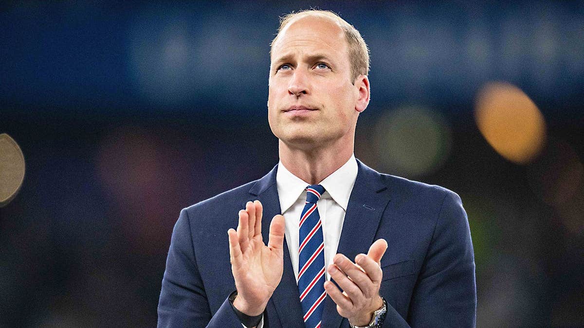 Prince William in a dark suit looking serious applauding from the field.