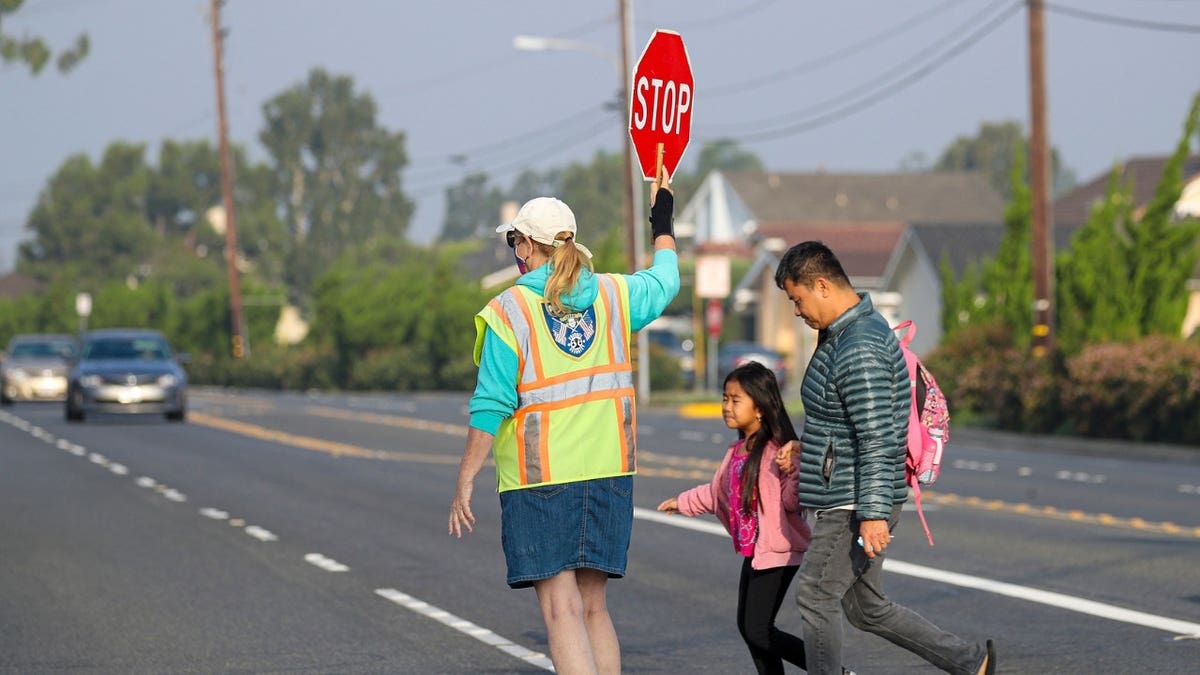 Un guardia de tráfico con un chaleco amarillo brillante ayuda a una niña vestida de rosa y a su padre a cruzar la calle