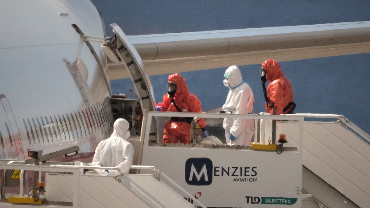 A passenger in a white protective suit boards a plane while officials in orange hazmat suits stand on the stairs.