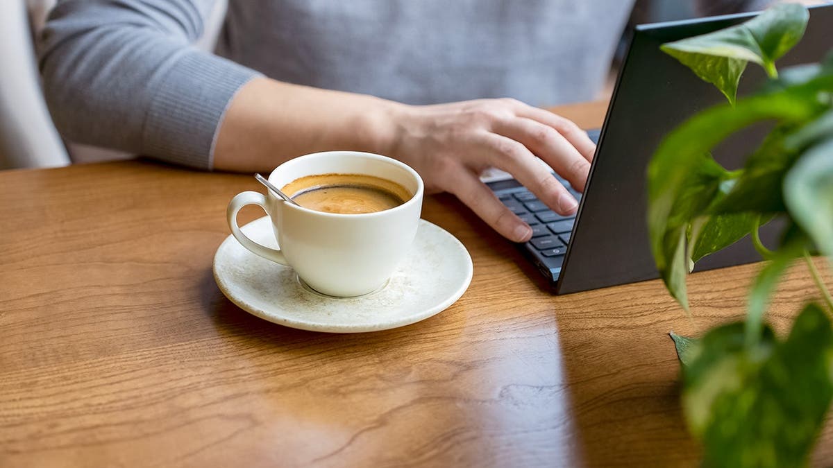 A person sits typing on a laptop with a cup of coffee beside him.