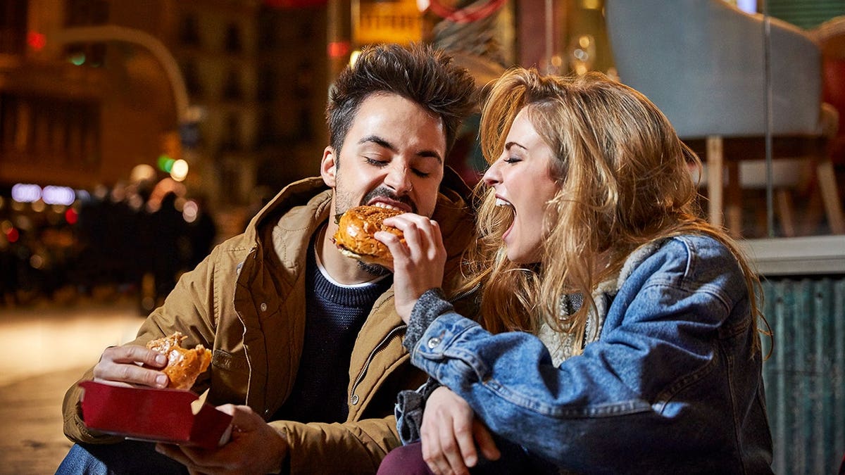 Cheerful woman feeding burger to man sitting outside restaurant at night