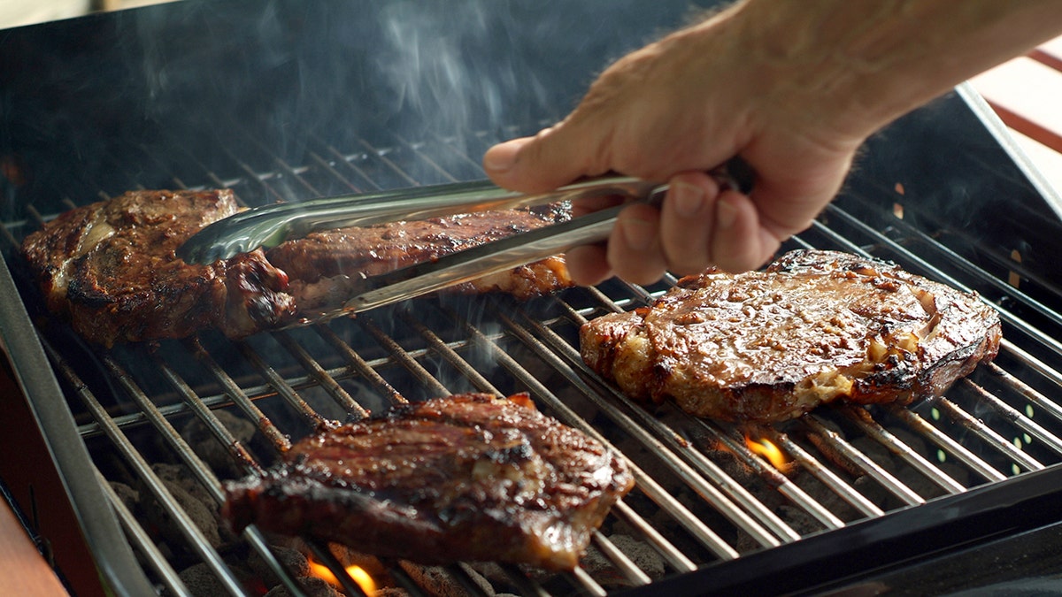 A man's hand holds tongs as he prepares to turn over a steak on the grill.