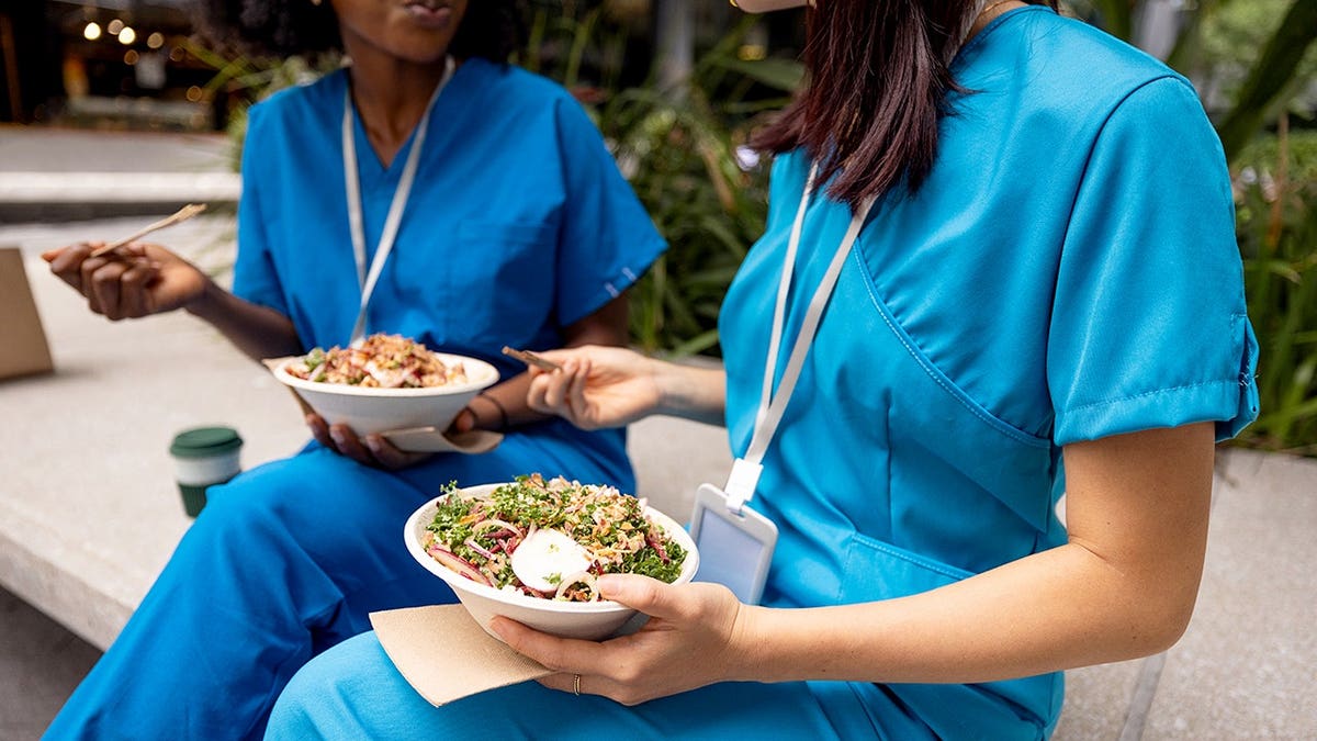 Two healthcare professionals in blue uniforms eating lunch outdoors.