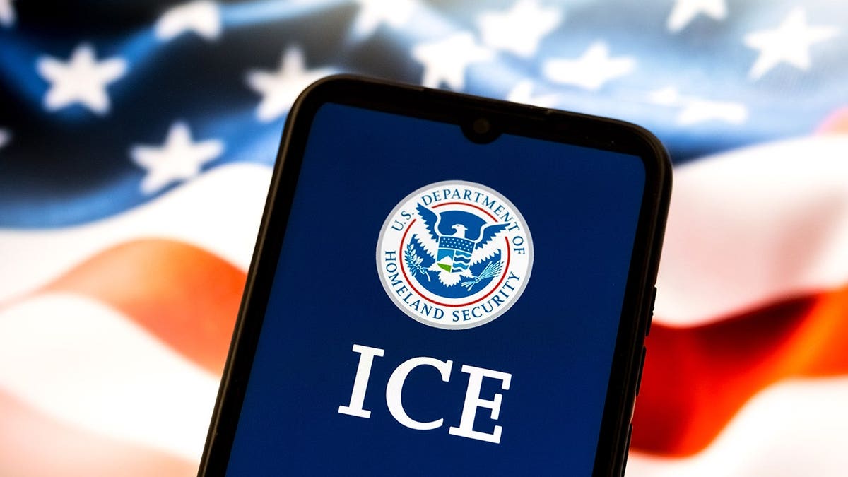 Smartphone showing US Department of Homeland Security and ICE with US flag on laptop screen in background