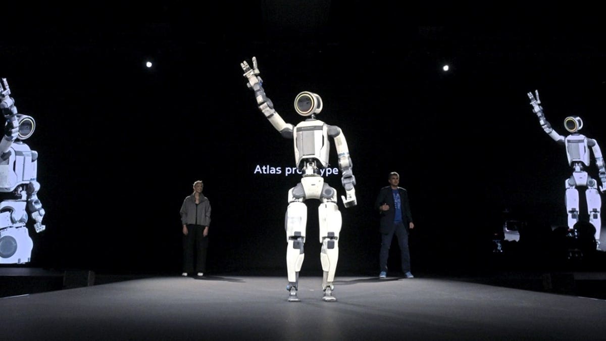 A Hyundai Atlas robot stands with an arm raised.