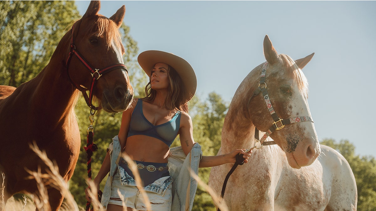 Jana Kramer posing with two horses while in a blue bra for her partnership with EBY.