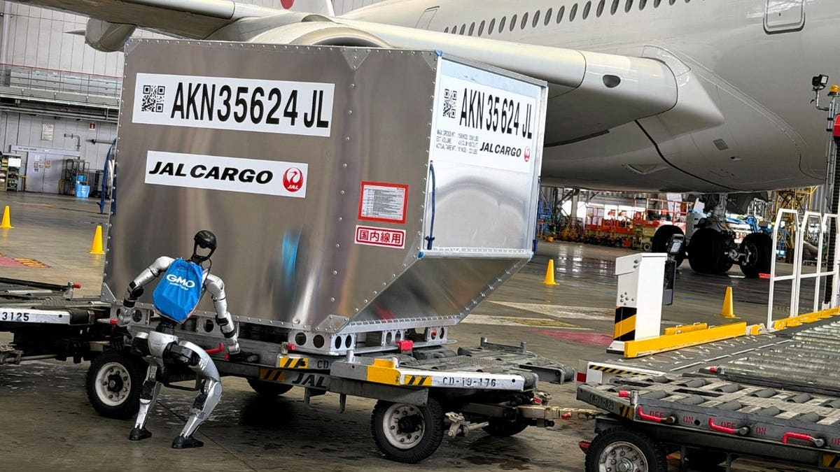 A humanoid robot bends over near a baggage cart.