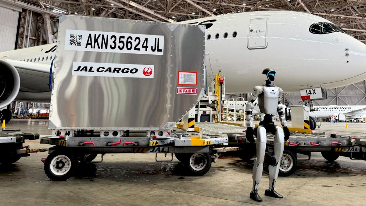 A robot stands in front of a baggage cart near a passenger airplane.