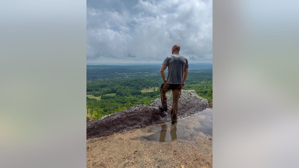 man stands on a cliff edge looking out over a scenic valley
