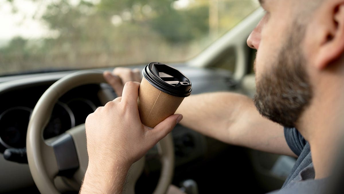 Bearded man driving while holding a paper coffee cup