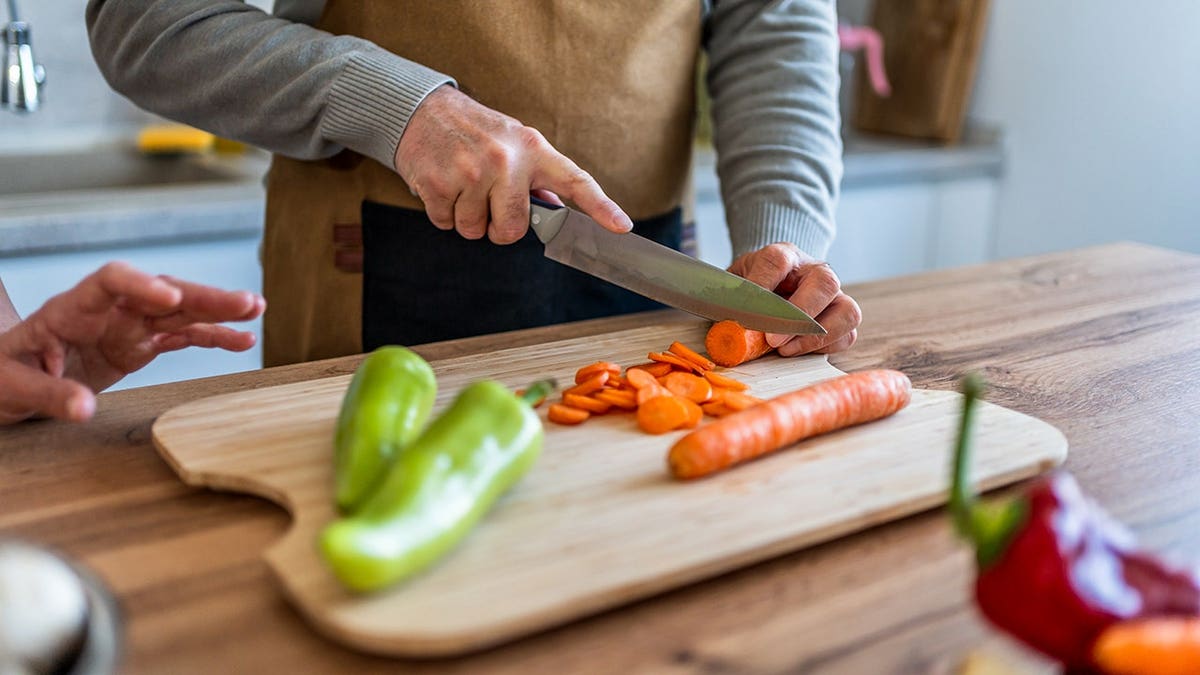 A mature man wearing an apron slicing carrots on a wooden cutting board in a kitchen
