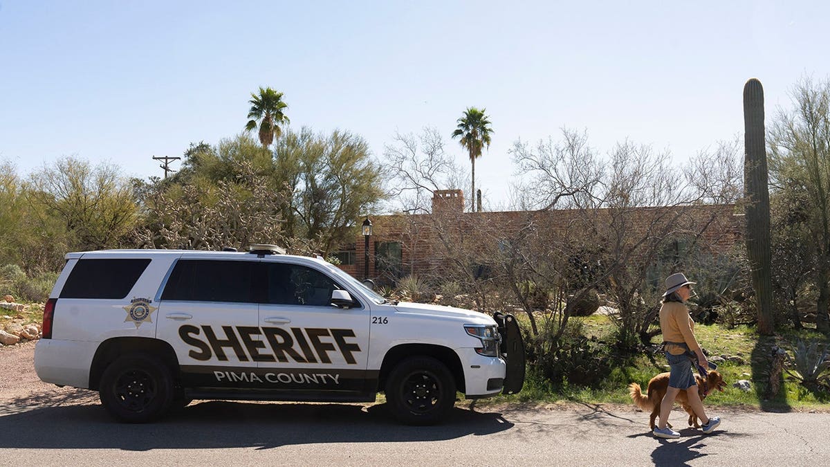 A woman walking her dog past a Pima County Sheriff's Office patrol car outside Nancy Guthrie's home in Tucson