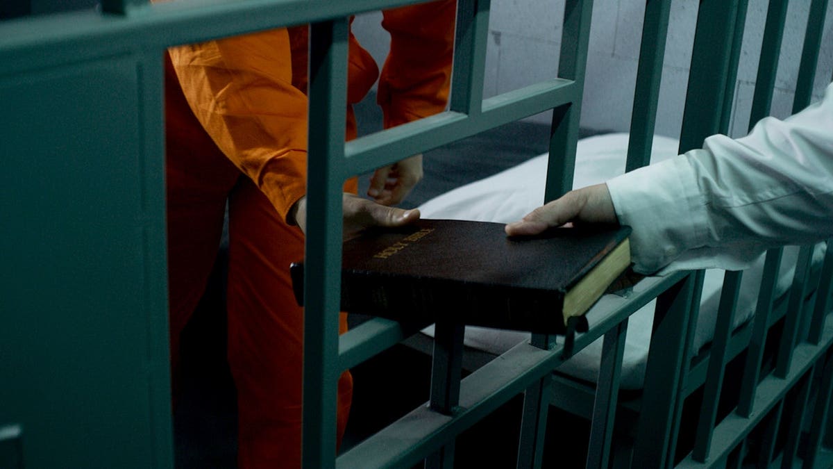 Prison officer handing Bible to male prisoner in orange uniform inside prison cell.