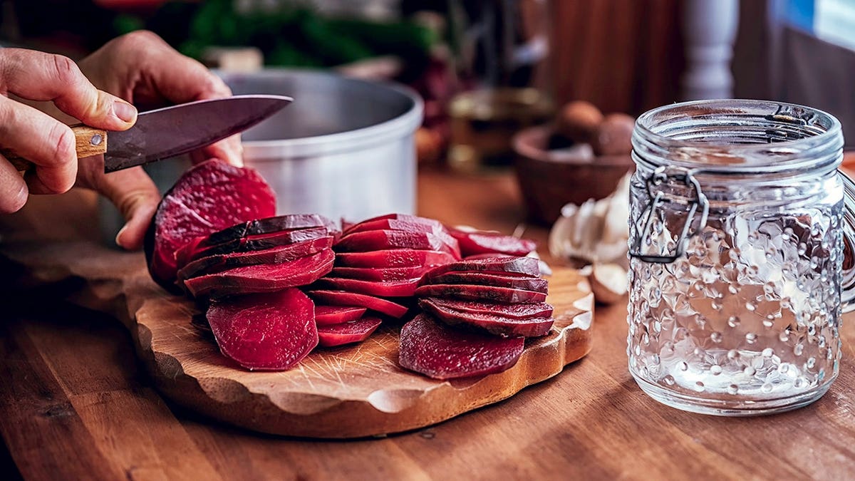 Man's hand slices red beets with a knife to be preserved in glass jar