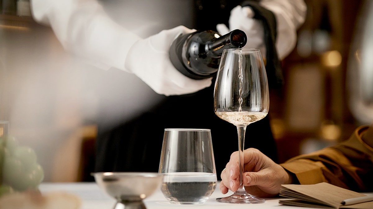 A sommelier pours white wine from a bottle into a long-stem glass while a man holds it.