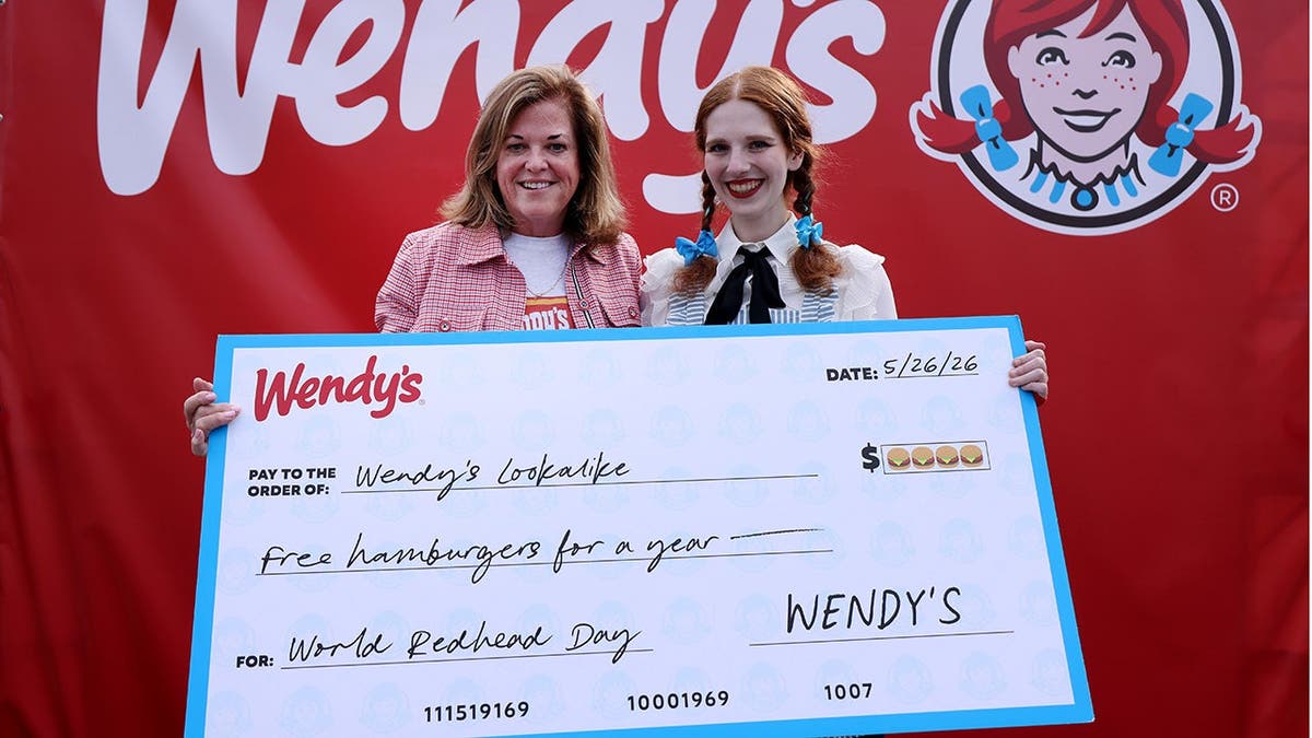A Wendy impersonator stands with another woman behind a large check written out for free hamburgers.