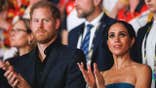 Prince Harry and Meghan Markle standing close to each other in an audience applauding and looking serious.