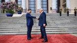 Xi Jinping and Donald Trump shake hands while standing on a red carpet in front of large stone steps.