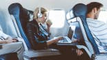 Young blonde woman sitting inside an airplane using a laptop and drinking coffee