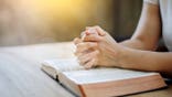 A woman praying with hands on an open Bible on a wooden table.