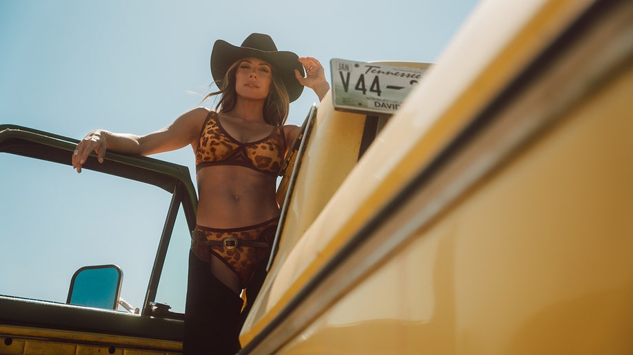 Jana Kramer standing on a pickup truck in a leopard-print underwear set and brown hat.