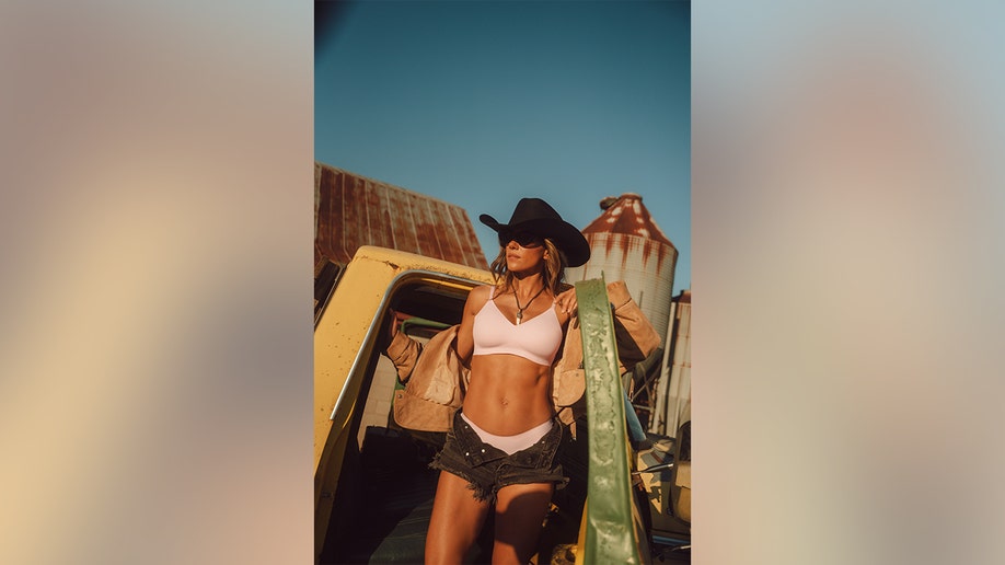 Jana Kramer posing in a pickup truck while in a pink bra and black shorts with a black cowboy hat.
