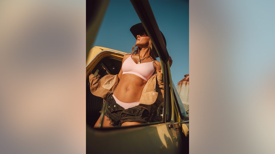 Jana Kramer looking off into the distance as she exists a pickup truck in a pink bra and black shorts in a campaign shoot.