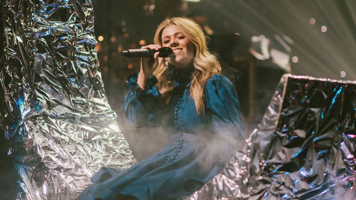 Anne Wilson usando um vestido azul sentada em uma meia-lua prateada cantando ao microfone.