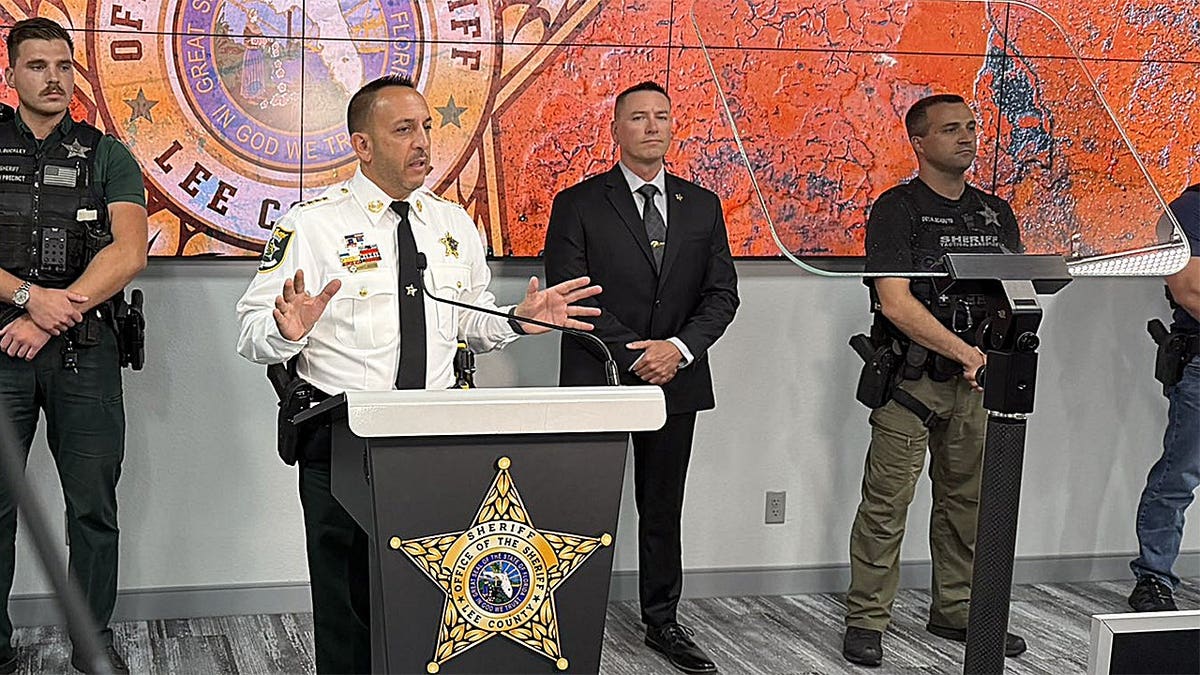 El sheriff del condado de Lee, Carmine Marceno, habla desde un atril durante una rueda de prensa, mientras los agentes e investigadores permanecen de pie detrás de él.