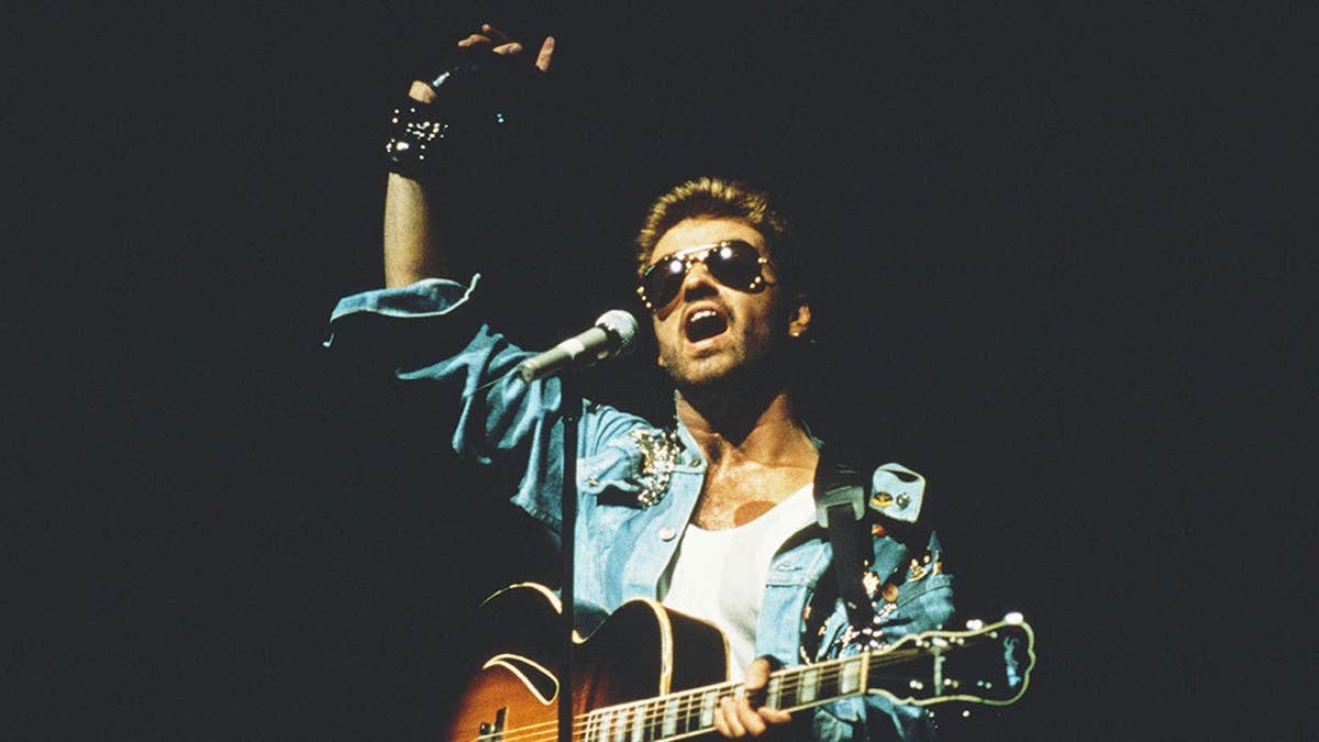 George Michael performing on stage wearing a leather jacket and aviator sunglasses.