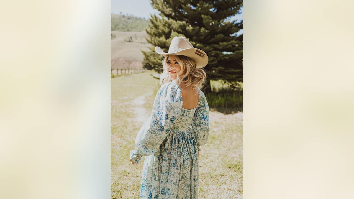 Anne Wilson usando um vestido branco e azul com um chapéu de cowboy sorrindo.
