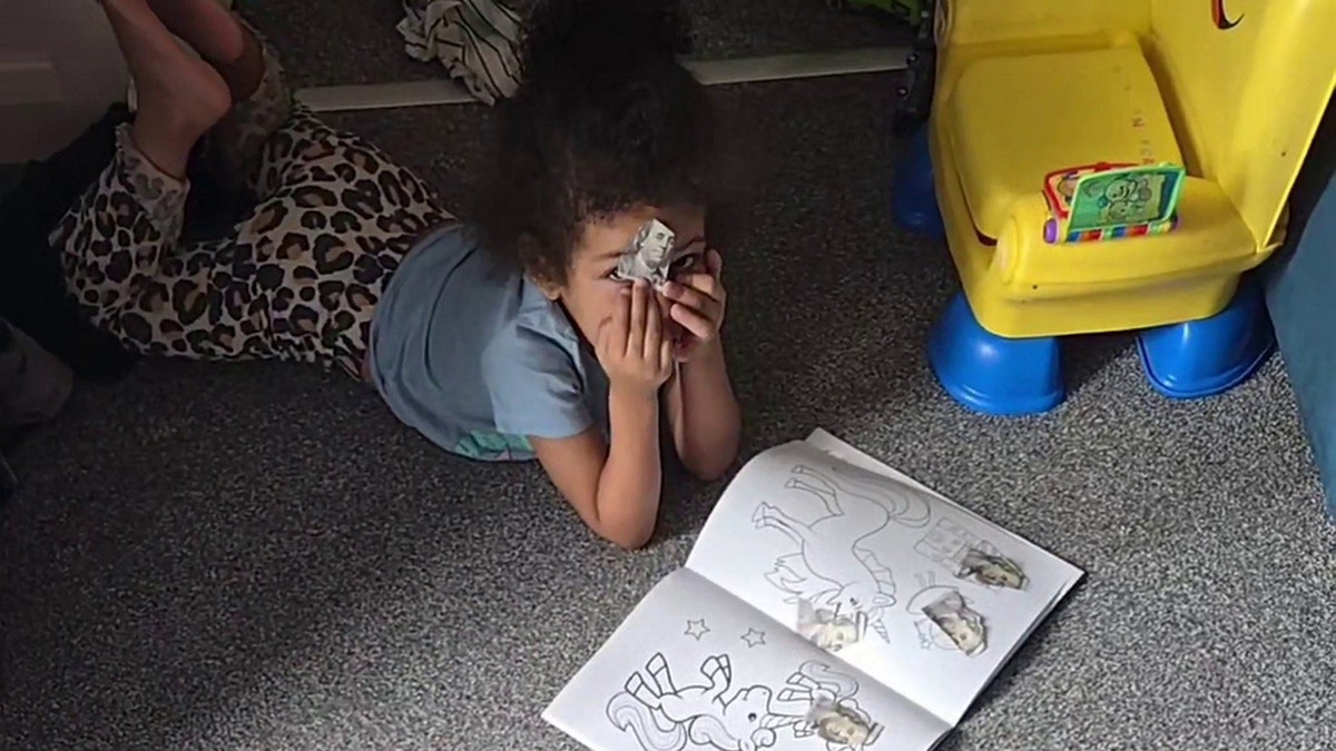 A girl puts a cut-up $100 bill over her face as she lays on the ground.