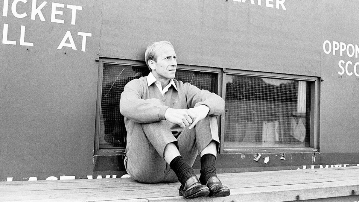 England footballer Bobby Charlton sitting at the base of a scoreboard playing cricket.