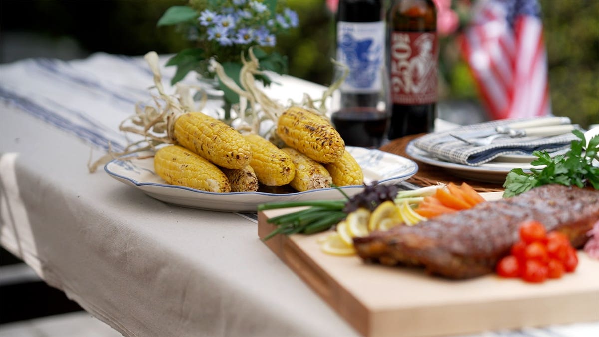 Corn on the cob sits on a table alongside grilled steak and fresh vegetables.
