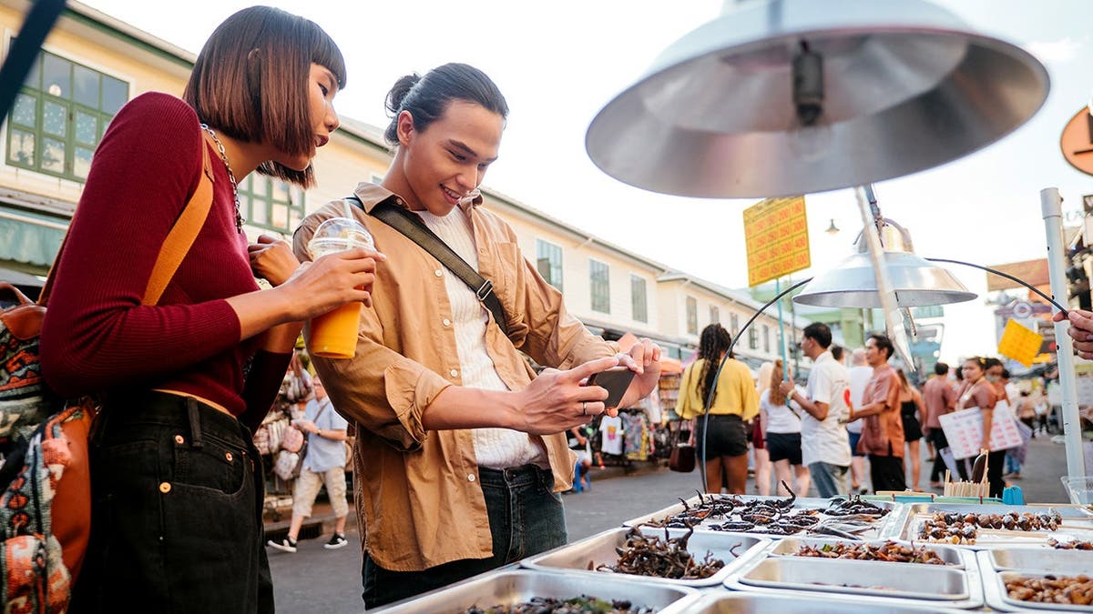 A couple looks at a selection of insect snacks from a food vendor. The man takes a picture of the display.